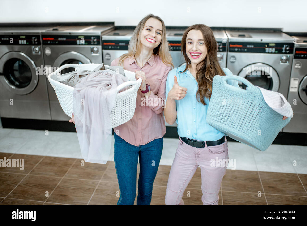 Portrait de deux amies gaies se tenant ensemble avec des paniers pleins de vêtements pour le lavage à la blanchisserie en libre-service Banque D'Images