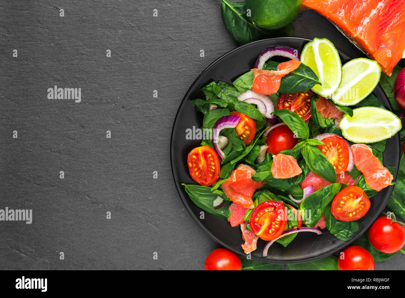 Alimentation saine. salade avec le saumon, les épinards, tomates cerises, oignons rouges, citron vert et le basilic dans la plaque noire sur fond d'ardoise noire. Une alimentation saine concept. Banque D'Images