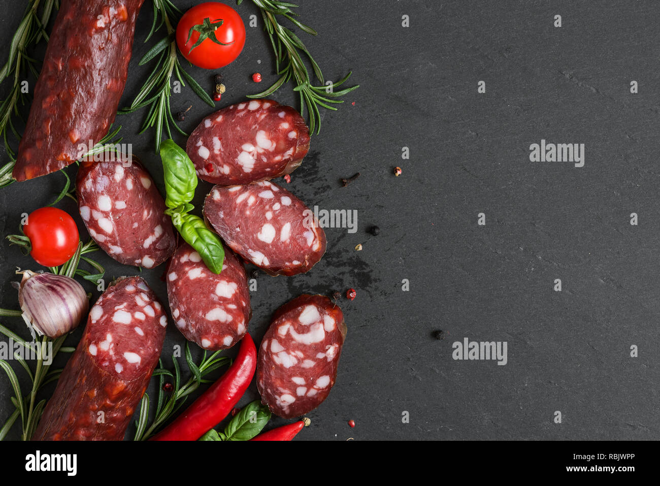 Tranches de salami fumé noir sur fond d'ardoise. Les saucisses avec le romarin, le basilic, la tomate et le poivre. top view with copy space Banque D'Images
