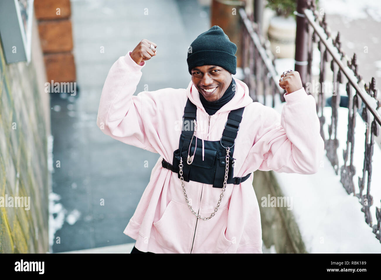 Style urbain élégant african american man en rose hoodie posée lors de l'hiver journée montre les muscles sur les mains. Banque D'Images