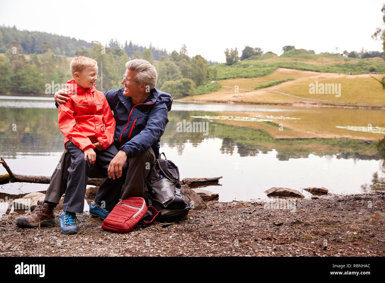 Petit-fils du grand-père assis sur son genou à la rive d'un lac smiling, Lake District, UK Banque D'Images