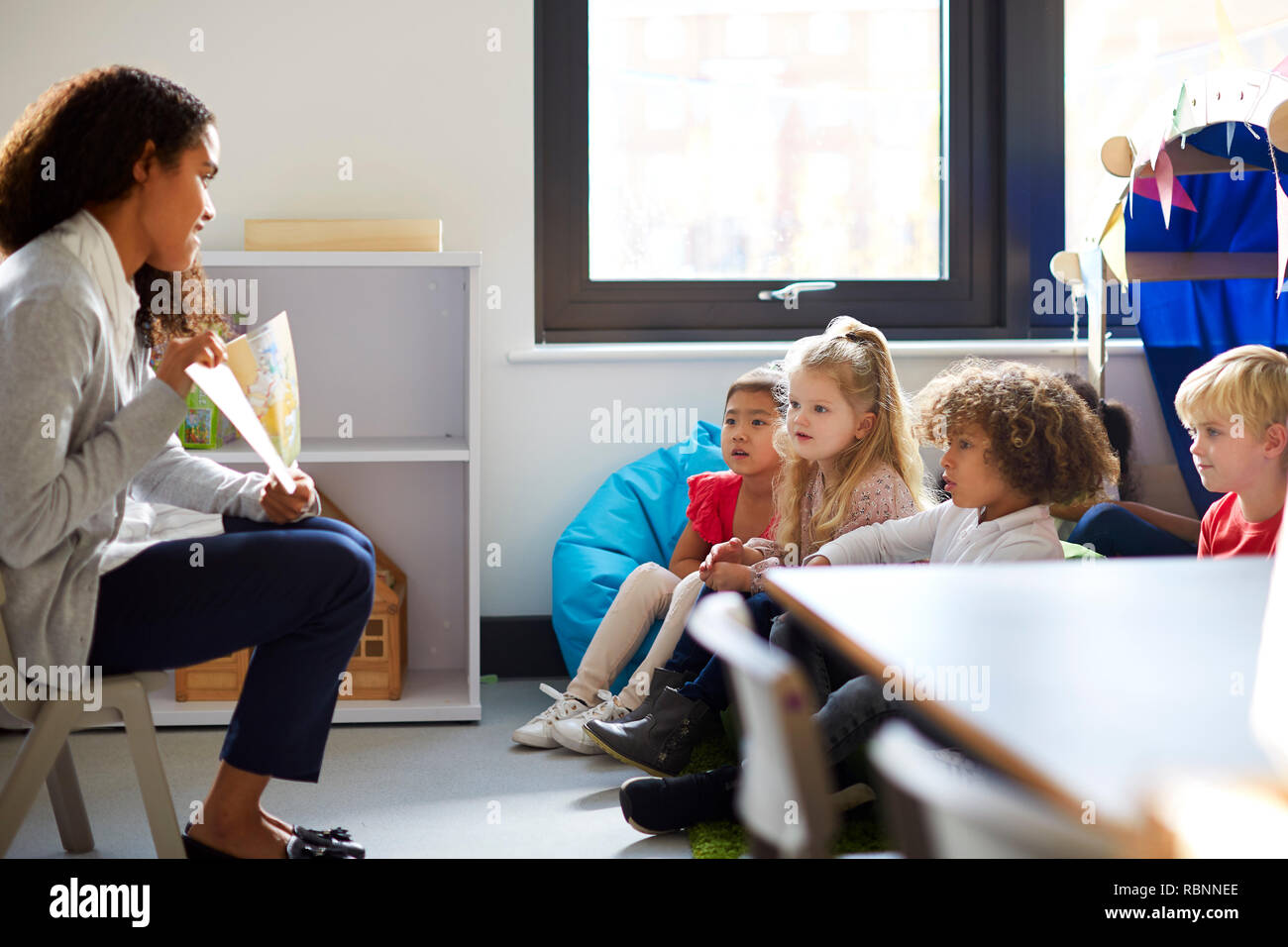 Vue latérale d'un enseignant de maternelle femme assise sur une chaise montrant un livre pour enfants dans une salle de classe Banque D'Images