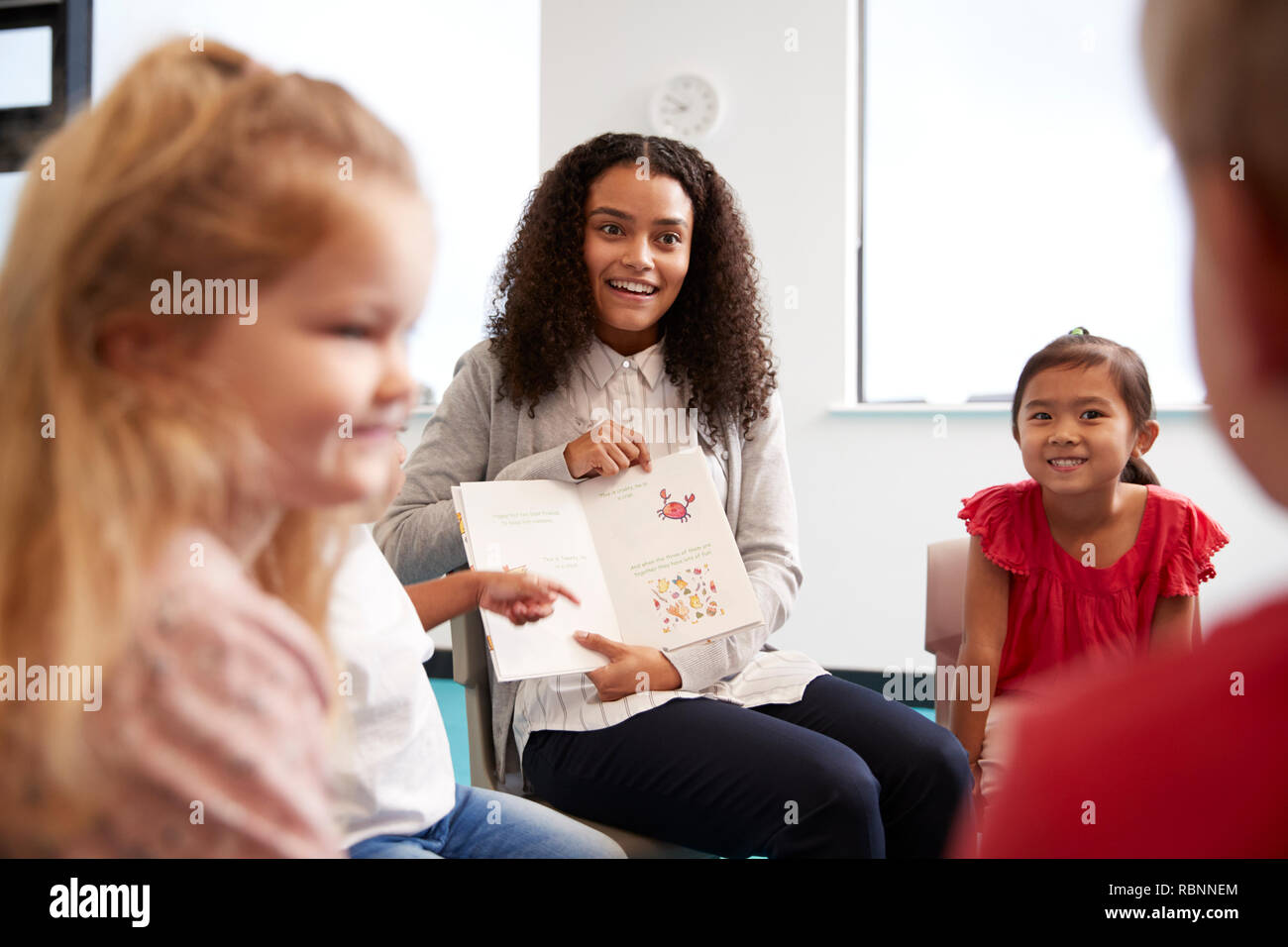 Plus d'épaule d'enseignante montrant une photo dans un livre d'un groupe d'enfants de maternelle assis sur des chaises dans une salle de classe, Close up Banque D'Images