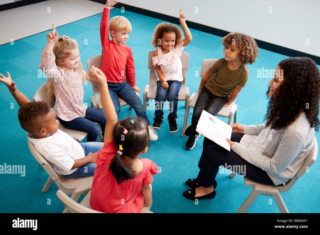 Jeune femme professeur de l'école de la lecture d'un livre à l'enfant Les enfants de l'école, assis sur des chaises en cercle dans la salle de classe la main pour répondre à une question, elevated view Banque D'Images