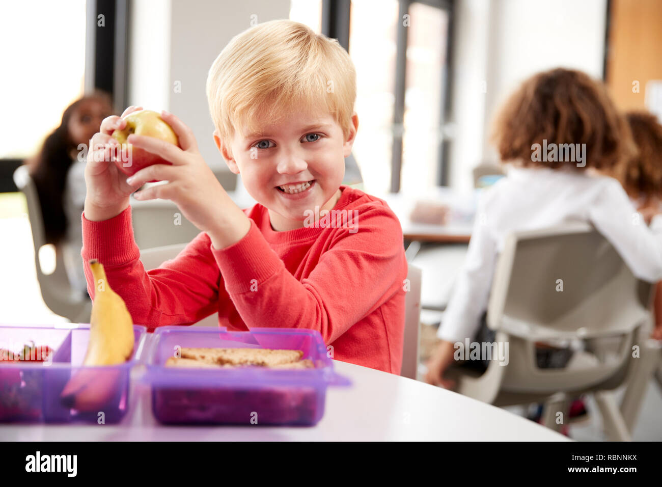 Jeune écolier blanc assis à une table en souriant et tenant une pomme dans une classe de maternelle pendant sa pause déjeuner, Close up Banque D'Images