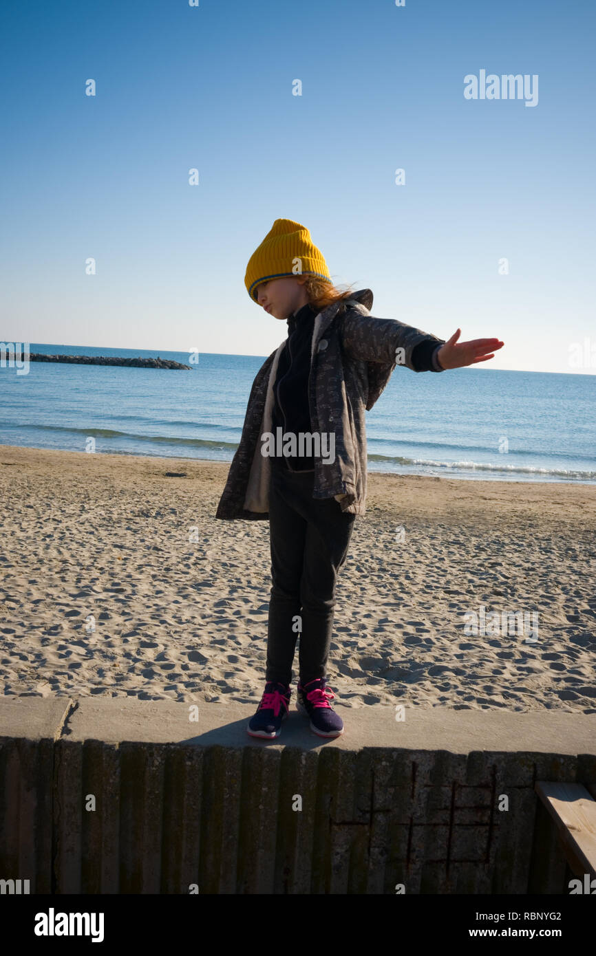 Une fillette de six ans avec yellow hat laineux et anorak se dresse sur une digue tout en faisant semblant de voler. Journée d'automne à Palavas les Flots, France Banque D'Images