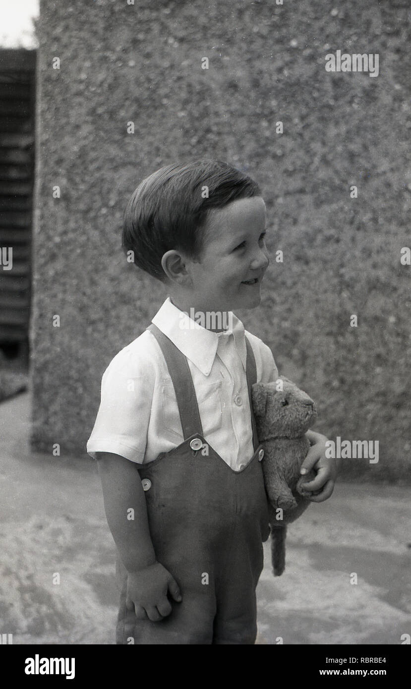 Années 1950, historiques, 'chauds' chappy...un jeune garçon avec son ours  en peluche et porter des shorts à bretelles, à l'autre manière et sourit  Photo Stock - Alamy