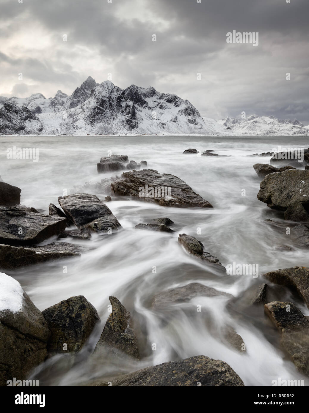 Paysage côtier en hiver avec le mouvement de l'eau entre de grandes pierres, dans l'arrière-plan d'une chaîne de montagnes avec de la neige, contraste élevé ciel - Lieu : Norvège Banque D'Images