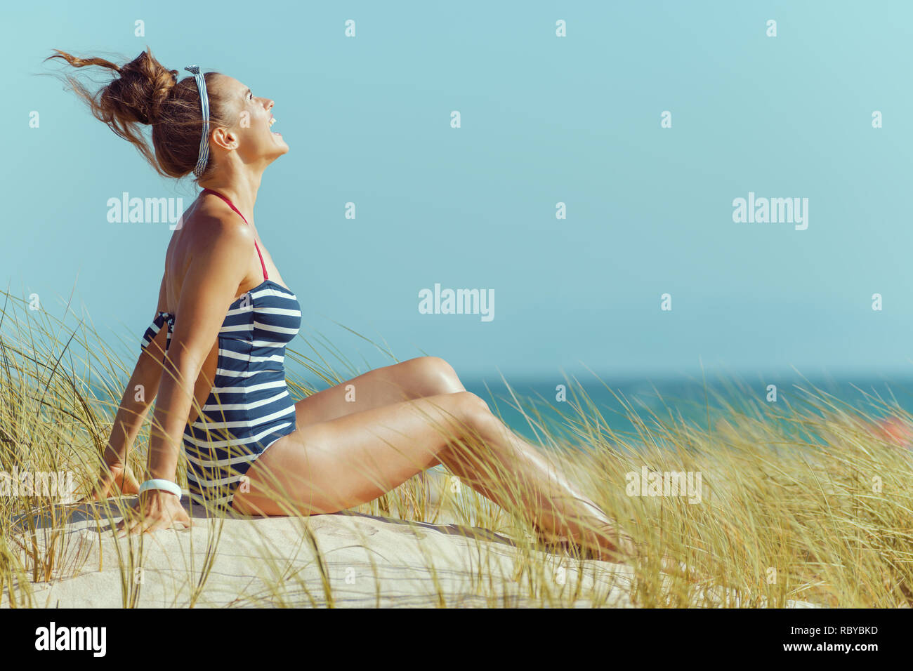 Smiling femme moderne en maillots à rayures sur le littoral reposant. bun coiffure. Midi d'été ensoleillé. ciel bleu. de protéger vos cheveux contre le soleil, la chaleur, une Banque D'Images