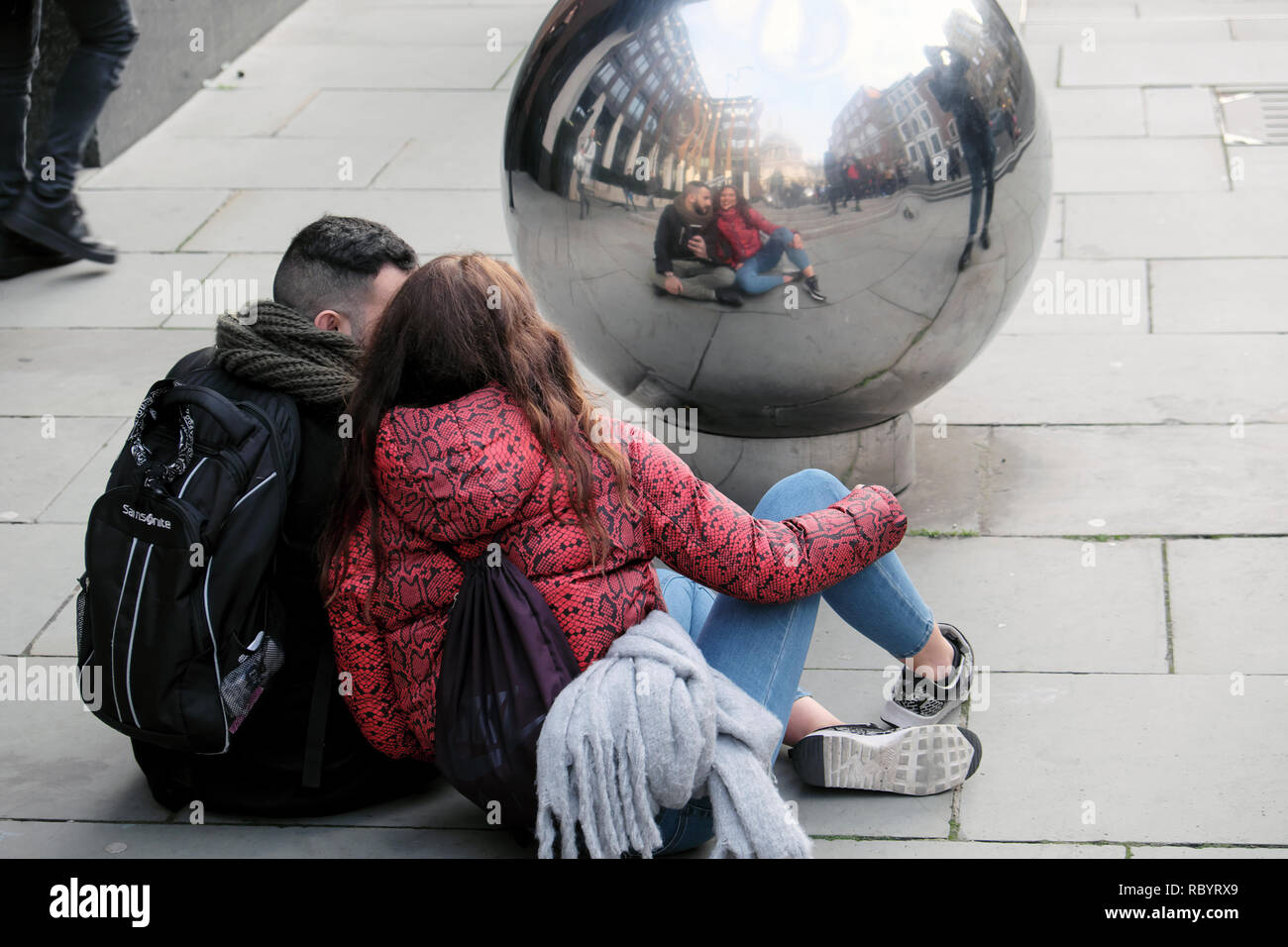 Jeune couple de touristes de prendre un avec un téléphone mobile selfies assis sur le trottoir à la recherche d'une balle d'argent en hiver London UK KATHY DEWITT Banque D'Images