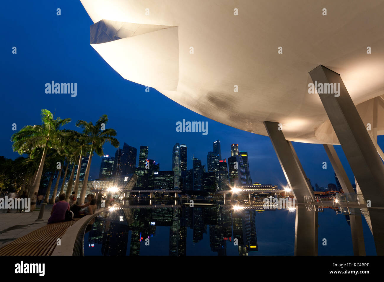 Allumé Musée ArtScience vu du dessous et des réflexions des gratte-ciel en CBD à Singapour au crépuscule. Plusieurs personnes assis et debout à côté de la piscine. Banque D'Images