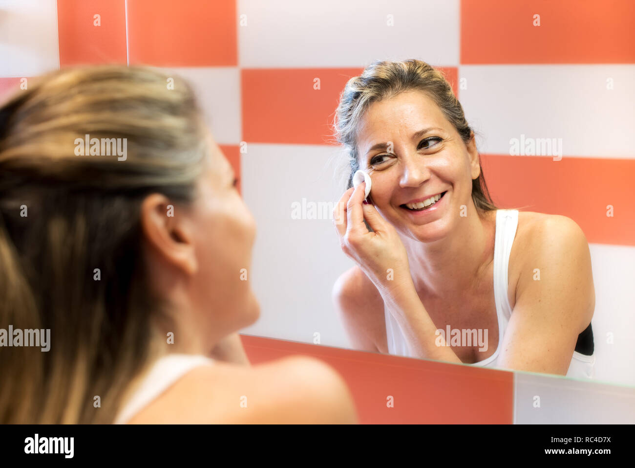 Smiling young blonde woman in white singlet dépose un miroir avec un coton, en regardant son reflet dans le miroir et de rire. Portrait buste de re Banque D'Images