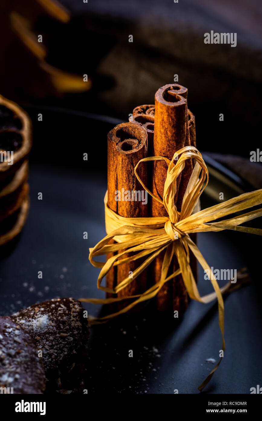 Photo verticale avec plusieurs bâtons de cannelle brun collée par cordon naturel. Bâtonnets sont sur la plaque noire avec des bonbons au chocolat et empilés anneaux orange à sec Banque D'Images