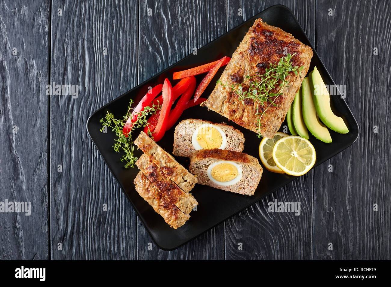 Le pain de viande avec oeuf dur dragées servi avec des tranches d'avocat et de poivron rouge sur un plateau rectangulaire noir sur une table de bois, vue de dessus, Fla. Banque D'Images