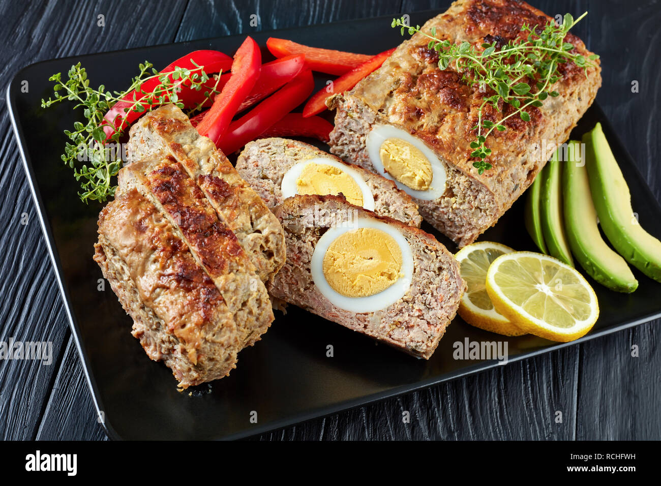 Viande farcies d'œufs servis avec le poivron rouge et les tranches d'avocat sur un plateau rectangulaire noir sur une table de bois, vue de dessus, close-up Banque D'Images