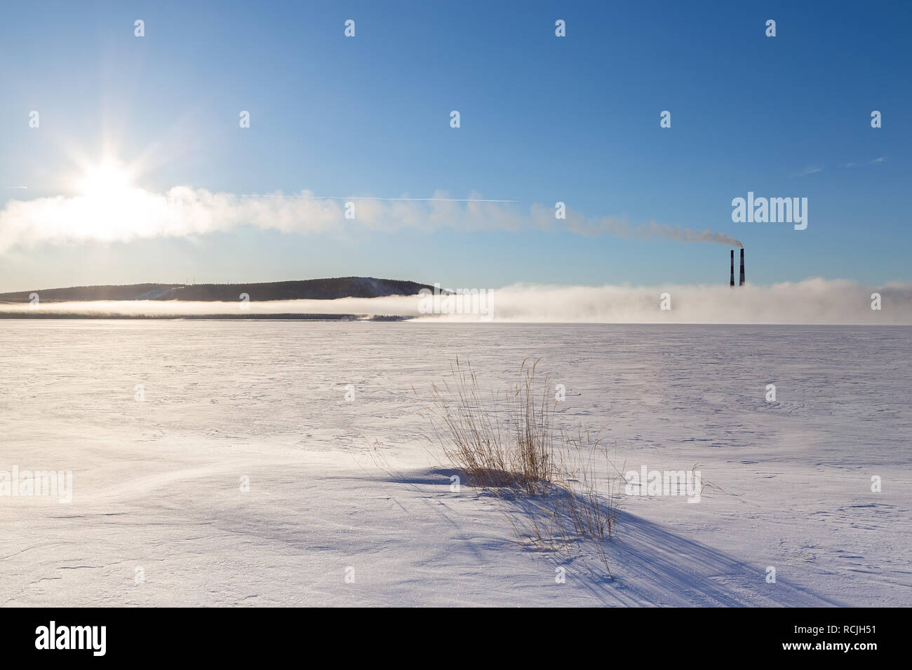 Panorama d'un lac gelé avec cheminées fumer dans le brouillard, le soleil et la piste de l'avion. Banque D'Images