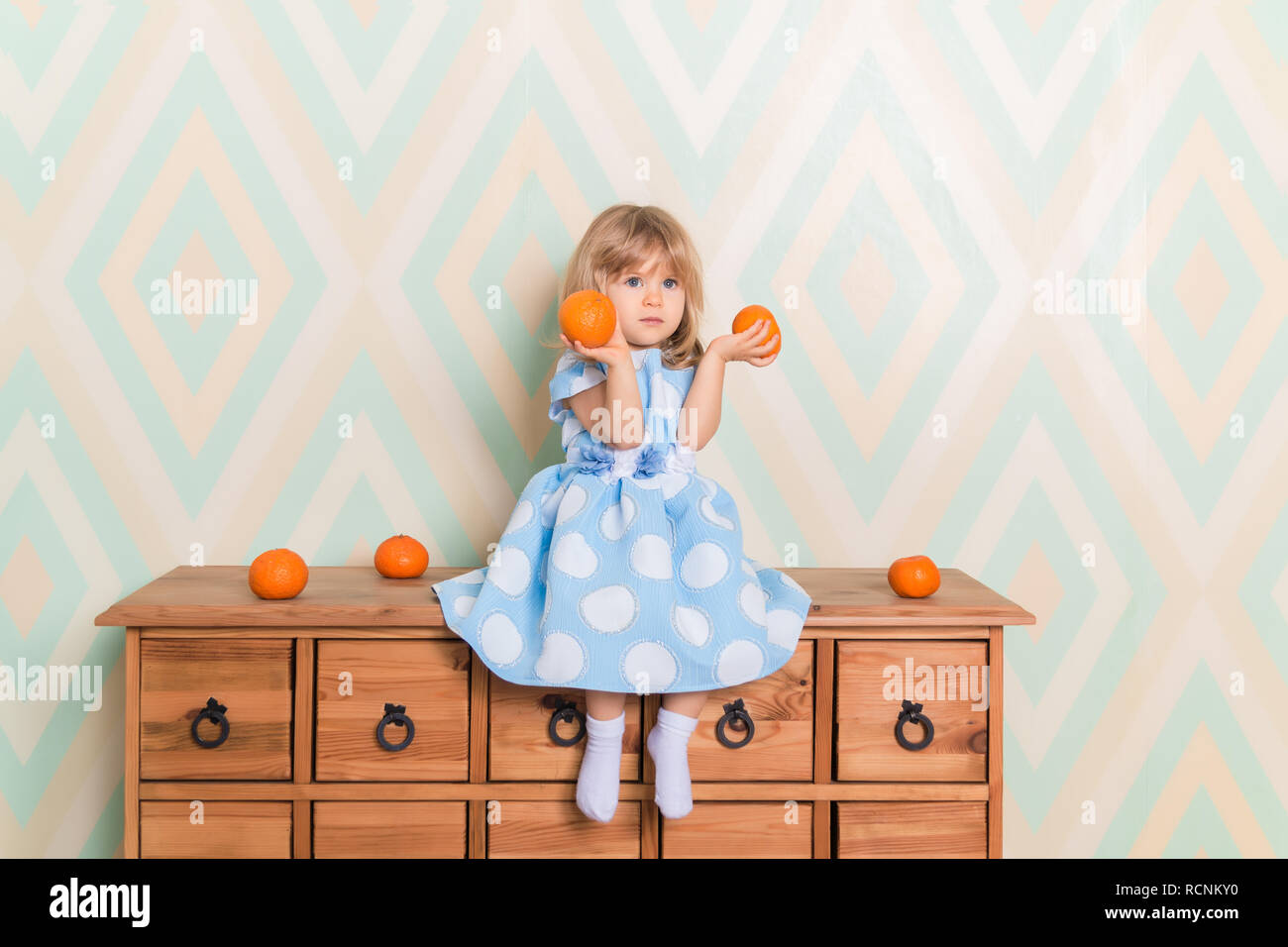 Tout-petit enfant bébé fille en robe bleu clair au sérieux assis sur le sol  en bois commode et holding fresh orange mandarine dans ses mains sur fond  de losange. Vacances d'hiver ou