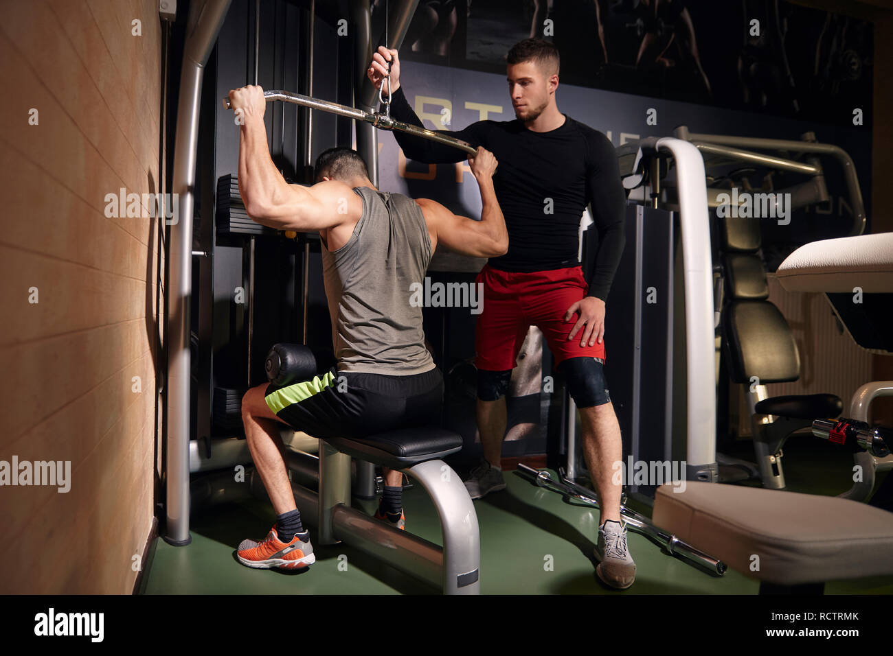 Deux jeune homme, 20-29 ans. Un homme d'exercer les muscles du dos, la formation sur lat pulldown machine, alors que d'autres ami ou d'un entraîneur est de l'aider. Dans une salle de sport, d'esprit Banque D'Images