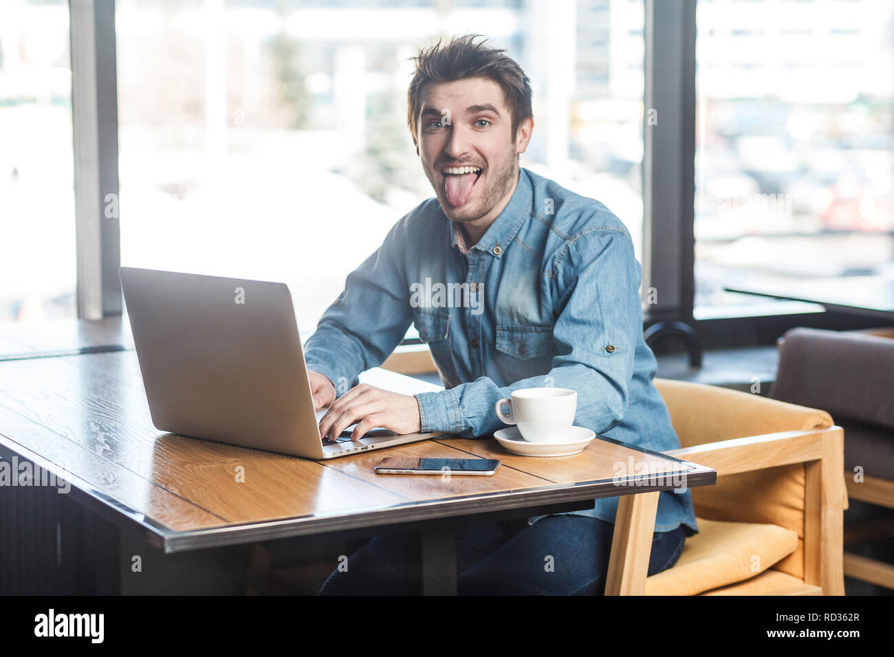 Le bouffon. Beau Portrait de jeunes barbus positive enfantin freelancer dans blue jeans chemise sont assis dans un café et travailler sur ordinateur portable montrant Banque D'Images Le bouffon. Beau Portrait de jeunes barbus positive enfantin freelancer dans blue jeans chemise sont assis dans un café et travailler sur ordinateur portable montrant Banque D'Images