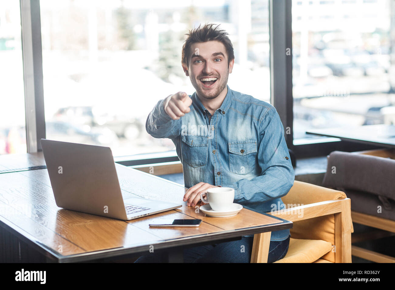 Hey you ! Portrait of happy young barbu positive freelancer dans blue jeans chemise sont assis dans un café et travailler sur ordinateur portable avec sourire à pleines dents et point Banque D'Images Hey you ! Portrait of happy young barbu positive freelancer dans blue jeans chemise sont assis dans un café et travailler sur ordinateur portable avec sourire à pleines dents et point Banque D'Images
