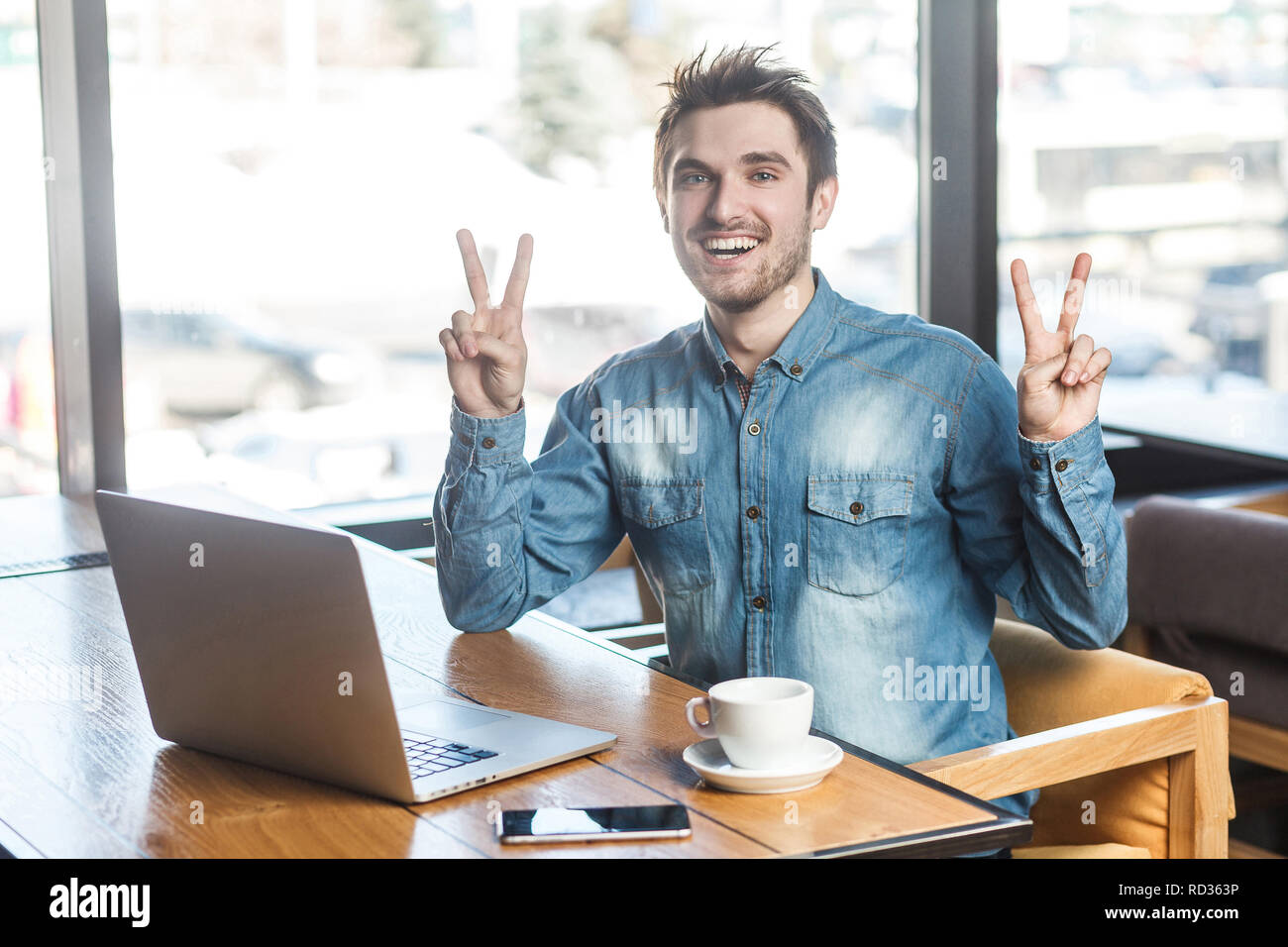 Beau Portrait de jeunes barbus positive succès pigiste en bleu jeans chemise sont assis dans un café et travailler sur ordinateur portable avec sourire et dentelée Banque D'Images Beau Portrait de jeunes barbus positive succès pigiste en bleu jeans chemise sont assis dans un café et travailler sur ordinateur portable avec sourire et dentelée Banque D'Images