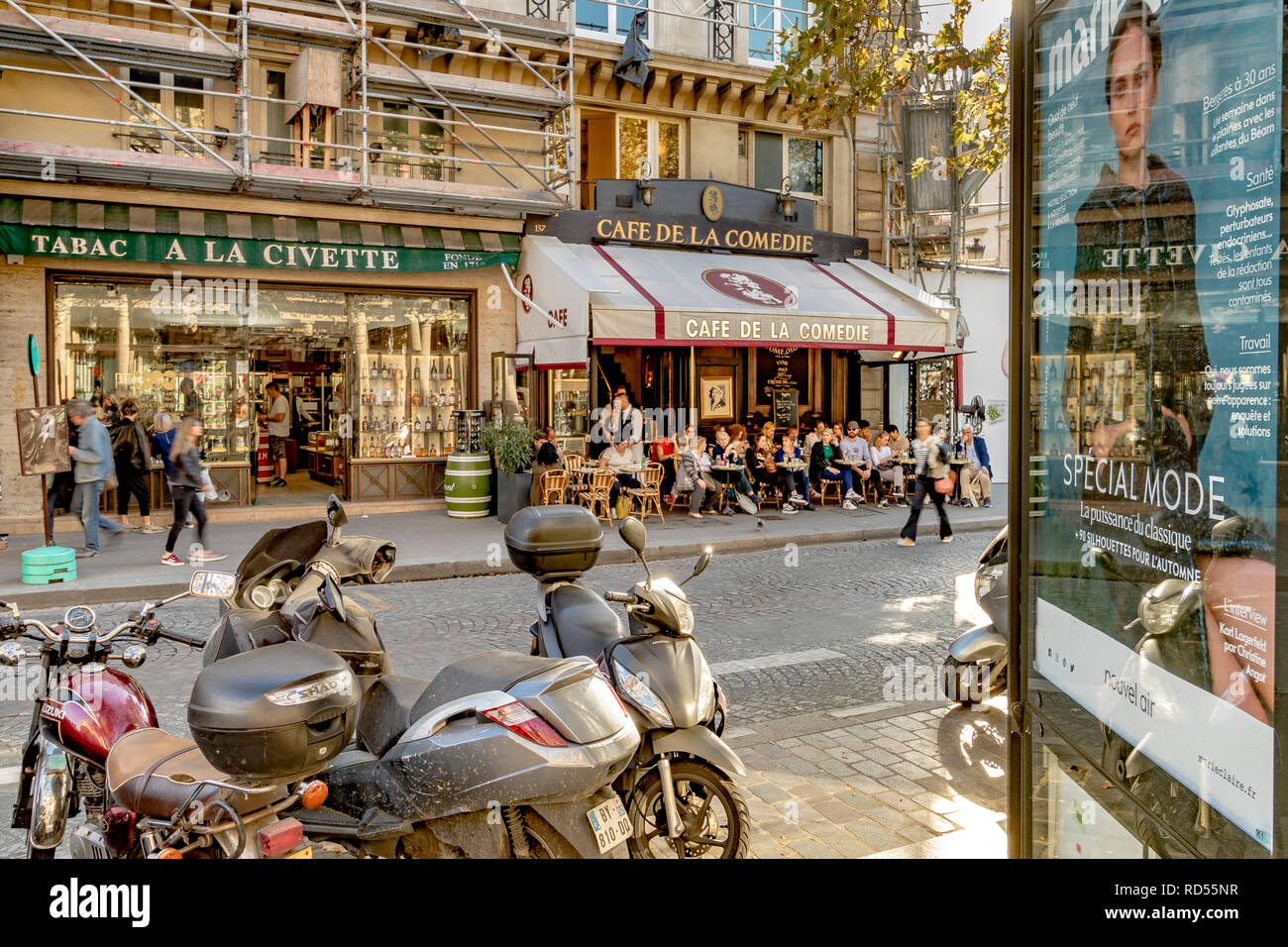 Des scooters garés dans une rue de Paris à côté d'un panneau publicitaire avec les gens assis dehors en train de déjeuner au café de la Comédie, rue Saint Honoré, Paris Banque D'Images