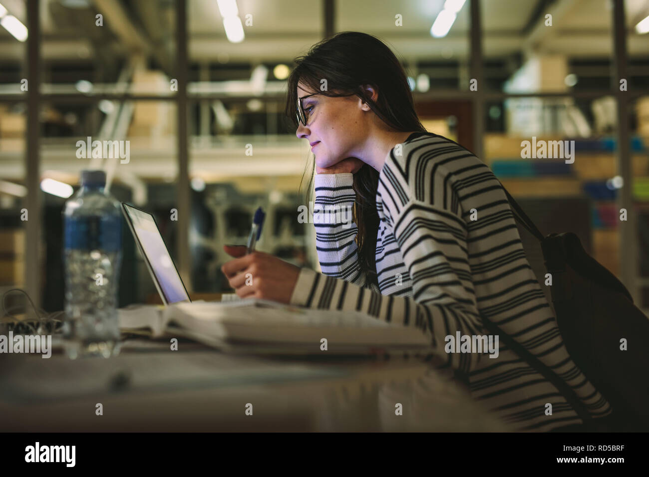 Étudiante à prendre des notes à partir d'un ordinateur portable à la bibliothèque. Young caucasian woman sitting at table faisant des affectations dans la bibliothèque du collège. Banque D'Images