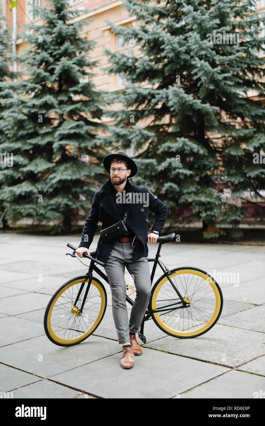 Portrait de beau barbu dans les tenues à l'écart et souriant tout en se penchant sur son vélo standing outdoors Banque D'Images