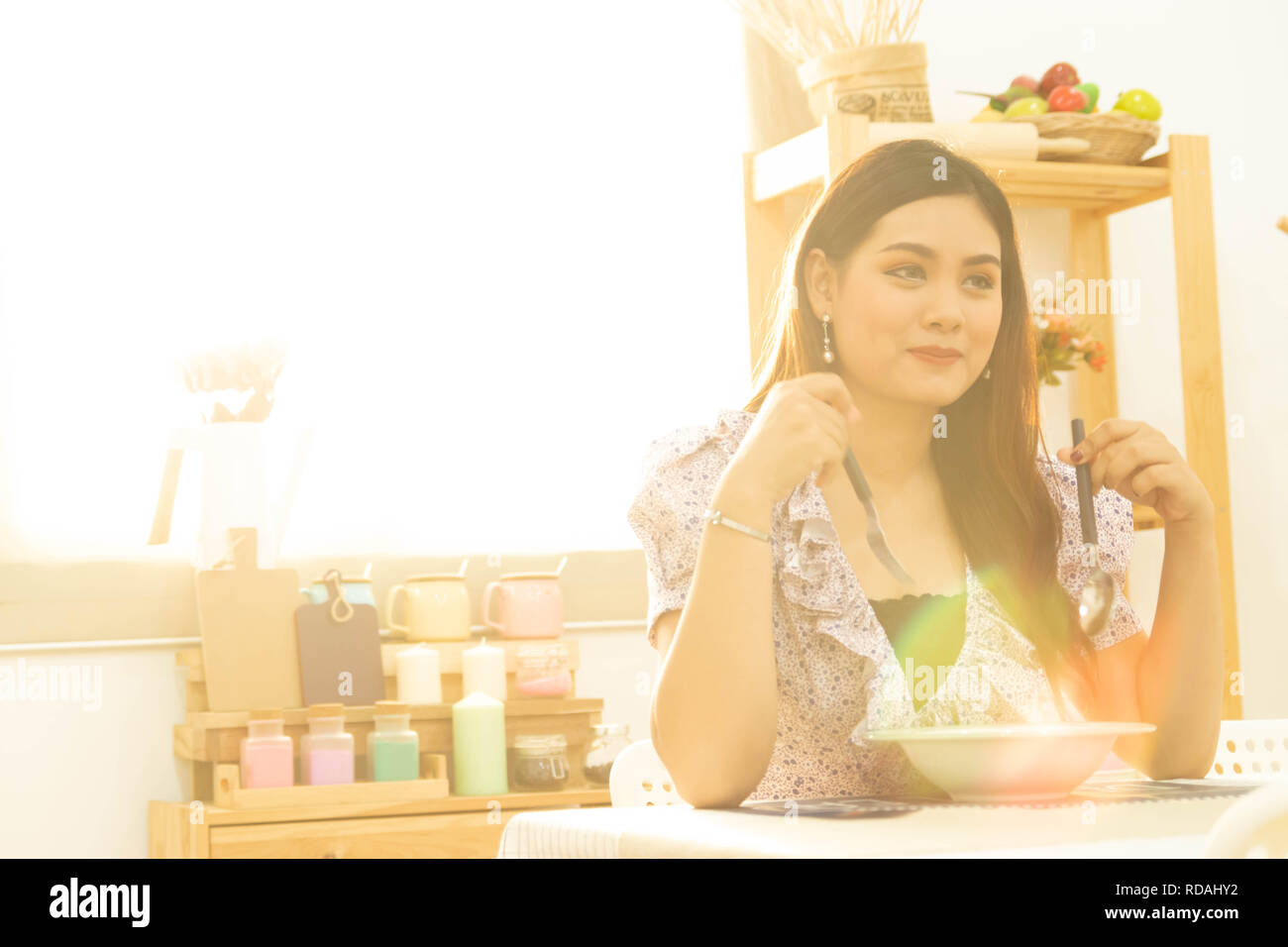 Jeune et heureux Asian woman eating healthy soup assis sur le tableau dans la cuisine avec une lumière chaude du soleil matinal with copy space Banque D'Images