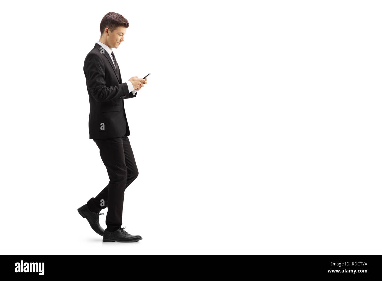 Profil de pleine longueur tourné d'un jeune homme dans un costume noir la marche et tapant sur un téléphone mobile isolé sur fond blanc Banque D'Images