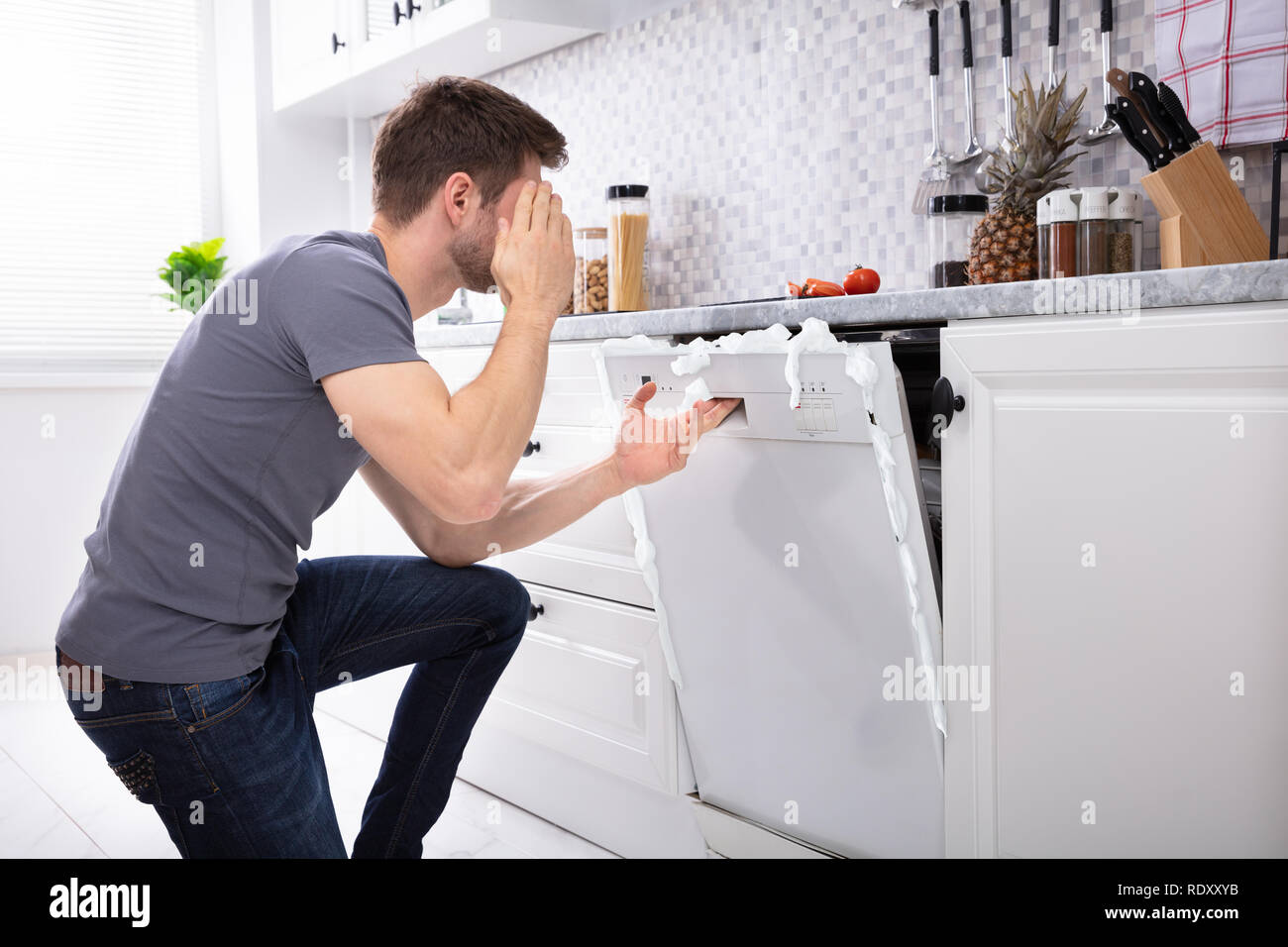 Contrarié homme assis en face de lave-vaisselle endommagé avec mousse venant de C Banque D'Images