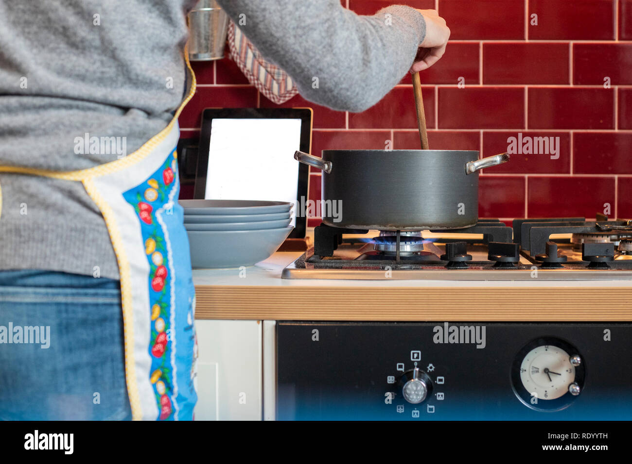 Femme avec tablier bleu, visage non visible, la cuisson dans un pot gris, d'une cuisine classique rouge, lors de la lecture de fiche sur tablette Banque D'Images