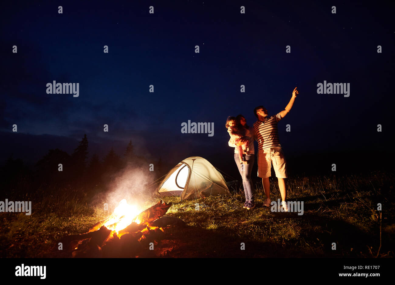 Camping familial dans les montagnes de nuit. Femme tenant des armes petite fille, homme pointant à bright stars dans ciel sombre en face de la tente et le brûlage de camp. Tourisme et de la beauté de la nature concept. Banque D'Images
