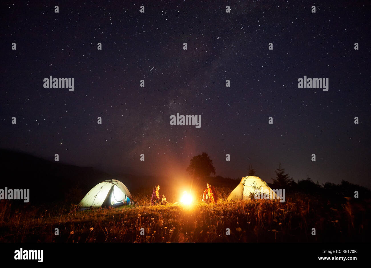 Nuit en camping dans les montagnes. Gravure de camp lumineux entre deux touristes, garçon et fille assise en face de l'autre en face de tentes lumineux bleu foncé sous un ciel étoilé sur les collines au loin. Banque D'Images