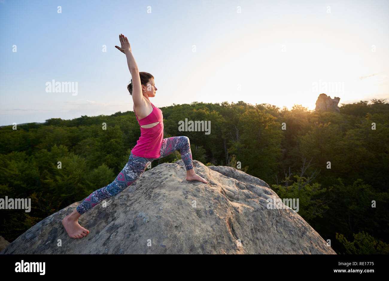 Belle slim woman doing stretching les exercices de yoga en haut du gros rocher sur green tree tops et ciel dégagé dans la matinée, l'arrière-plan. Le sport, yoga, fitness et d'un style de vie sain concept. Virabhadrasana Banque D'Images