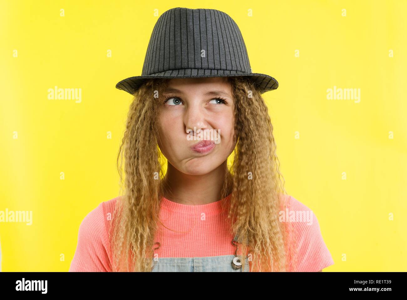 Jolie Fille 12-13 ans, blonde, aux cheveux bouclés dans un chapeau, l'air songeur de côté, en pensant à l'école. Concept d'expressions du visage, jaune backgrou Banque D'Images
