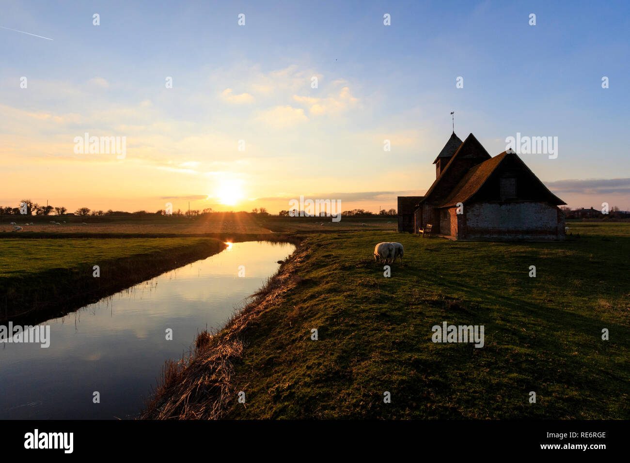 St Thomas Becket une église à Fairfield, dans le Kent en Romney Marsh. L'église en brique et bois isolés sur le terrain marécageux entouré par les moutons et les fossés au coucher du soleil, le coucher du soleil. Nuages dans le ciel, le soleil bas près de l'horizon. L'orange au bleu ciel. Banque D'Images