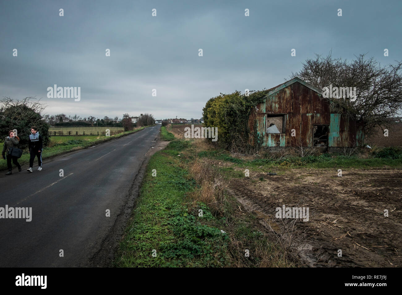 Deux adolescents à pied le long de la route de comté de bâtiments agricoles abandonnés passé Kent, UK Banque D'Images