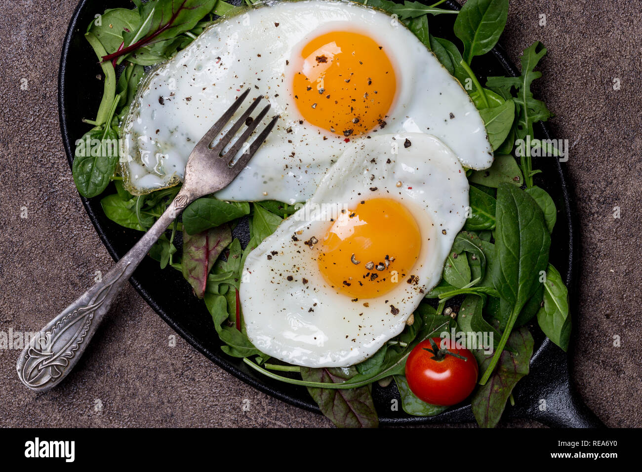Deux œufs frits sur poêle avec roquette, épinards et tomates cerise, avec la fourchette à fond brun, concept d'un petit-déjeuner sain, Close up, macro. Haut de la vie Banque D'Images Deux œufs frits sur poêle avec roquette, épinards et tomates cerise, avec la fourchette à fond brun, concept d'un petit-déjeuner sain, Close up, macro. Haut de la vie Banque D'Images