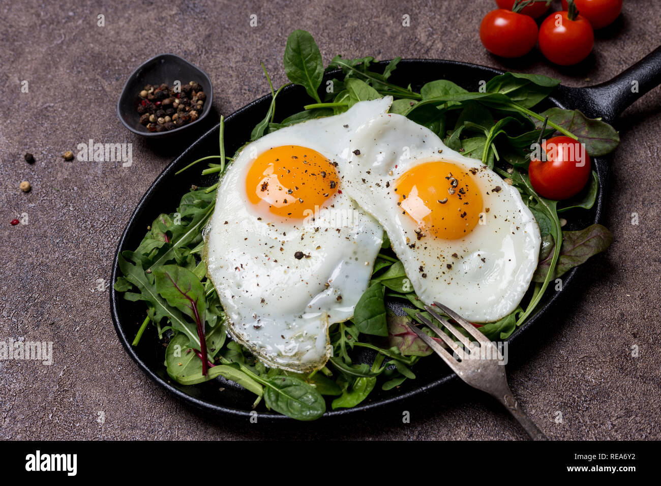 Deux œufs frits, Sunny Side Up, le pan avec des herbes, roquette, épinards, tomates cerise et poivre, avec une fourchette d'un fond brun. Concept de vie sain Banque D'Images Deux œufs frits, Sunny Side Up, le pan avec des herbes, roquette, épinards, tomates cerise et poivre, avec une fourchette d'un fond brun. Concept de vie sain Banque D'Images