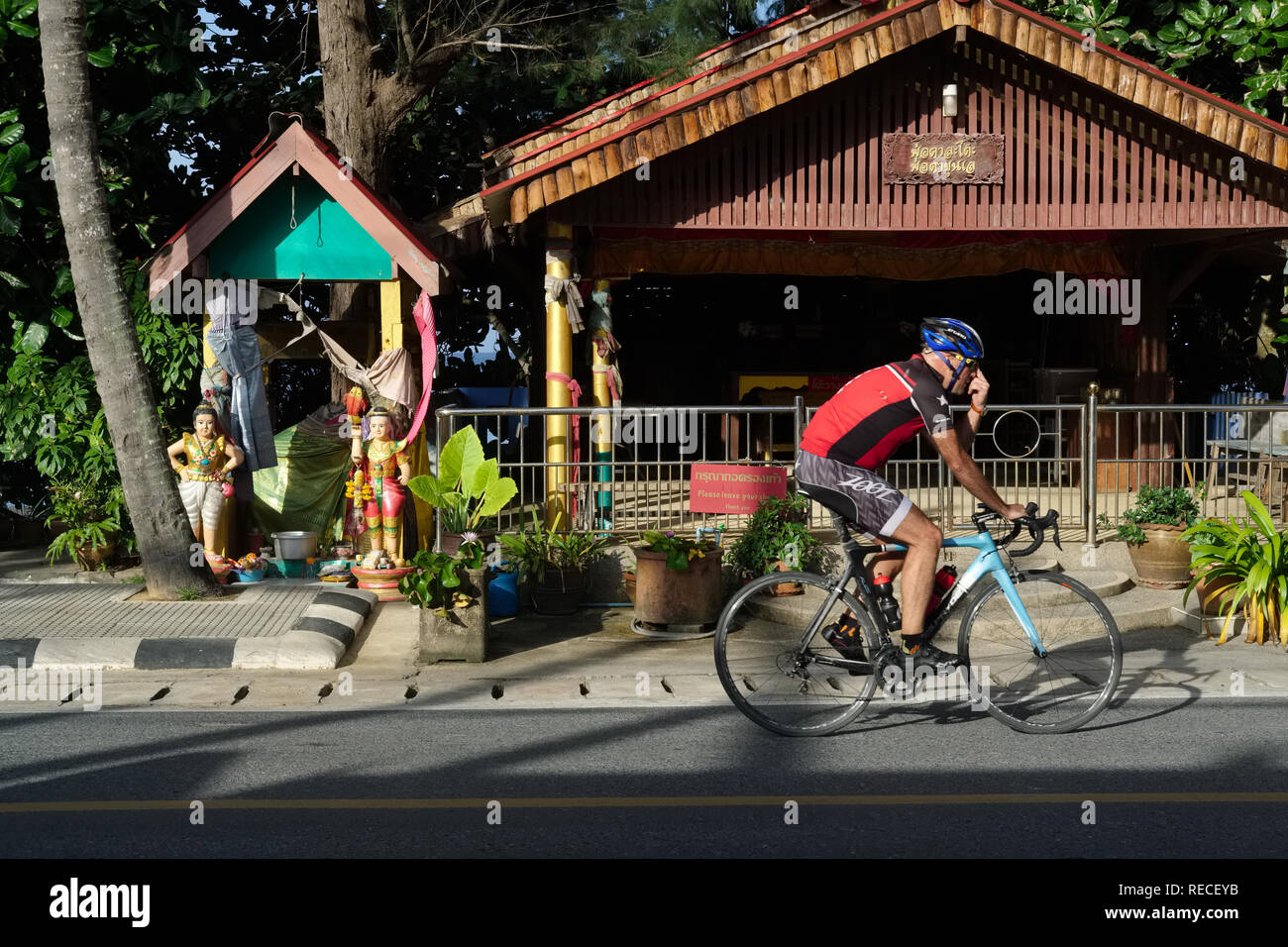 Un cycliste de l'équitation à travers Nai Thon, Phuket, Thaïlande ; en arrière-plan à gauche une prière de culte à une déesse de la mer ; plus de retard sur plage de Nai Thon Banque D'Images