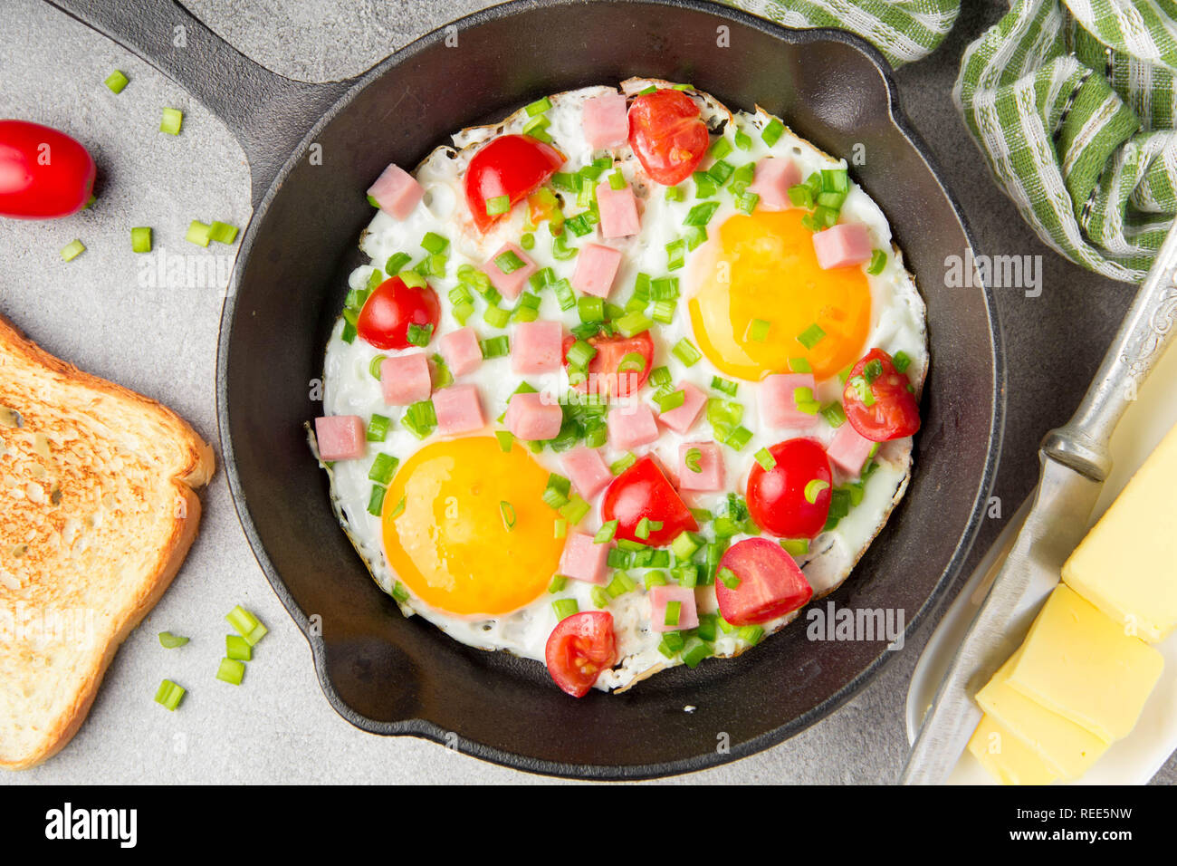 Oeufs frits en noir poêle avec du jambon, tomates cerises et oignons verts de printemps. Délicieux petit déjeuner avec du pain grillé et du beurre. Le classique Anglais Banque D'Images