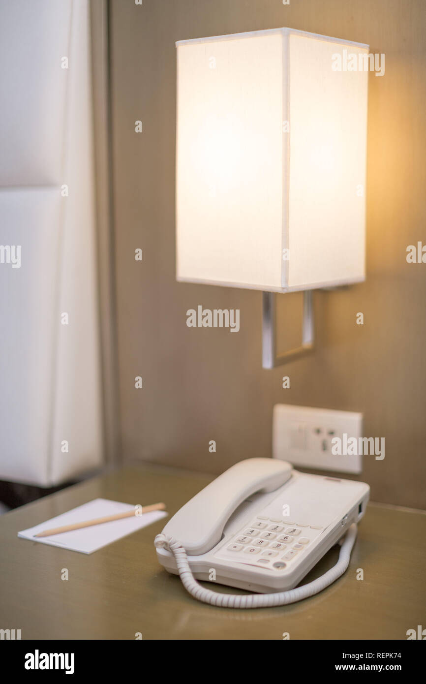 Chambre de l'hôtel téléphone blanc sur une table de chevet avec notes et  crayon une lampe de chevet et la prise de l'appareil le tout dans une scène  lumineuse Photo Stock -