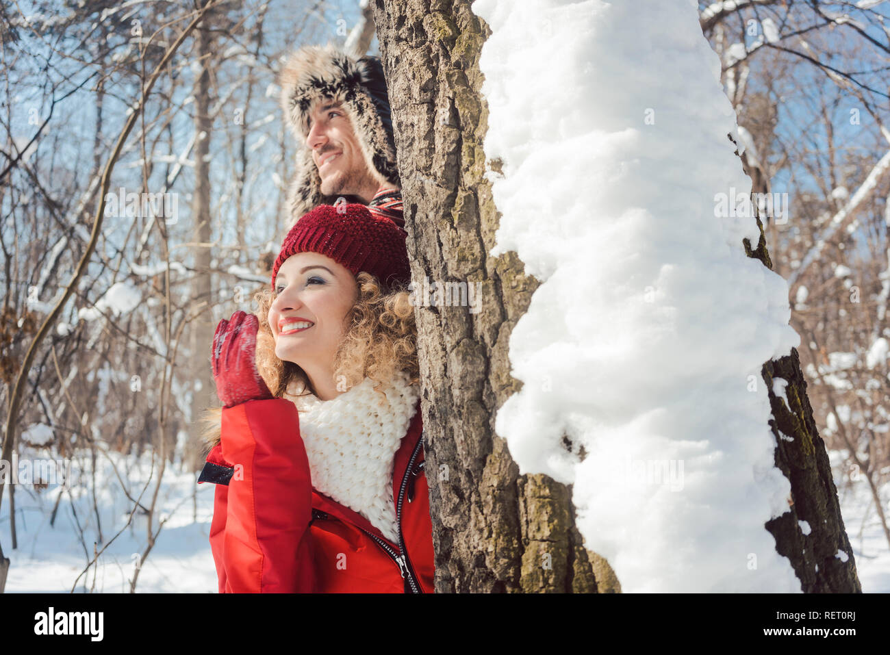 Playful couple se cacher derrière un tronc d'arbre dans la neige Banque D'Images