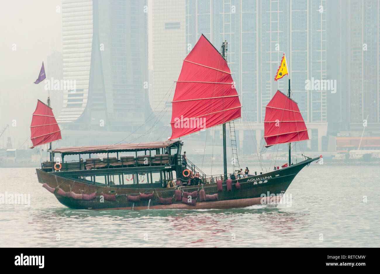 Bateau à voile avec voiles rouge et jaune et violet drapeaux, avec personnes à bord, dans le port de Hong Kong brumeux, la Chine. Banque D'Images