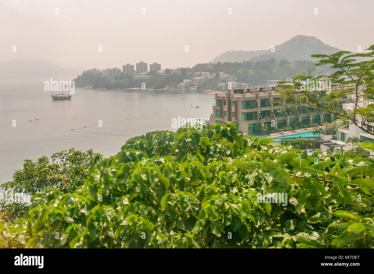 Feuilles vertes sur les arbres abondent dans ce point de vue surplombant Ocean avec voile sur l'eau à Hong Kong, Chine, le jour brumeux dans ville d'Asie. Banque D'Images