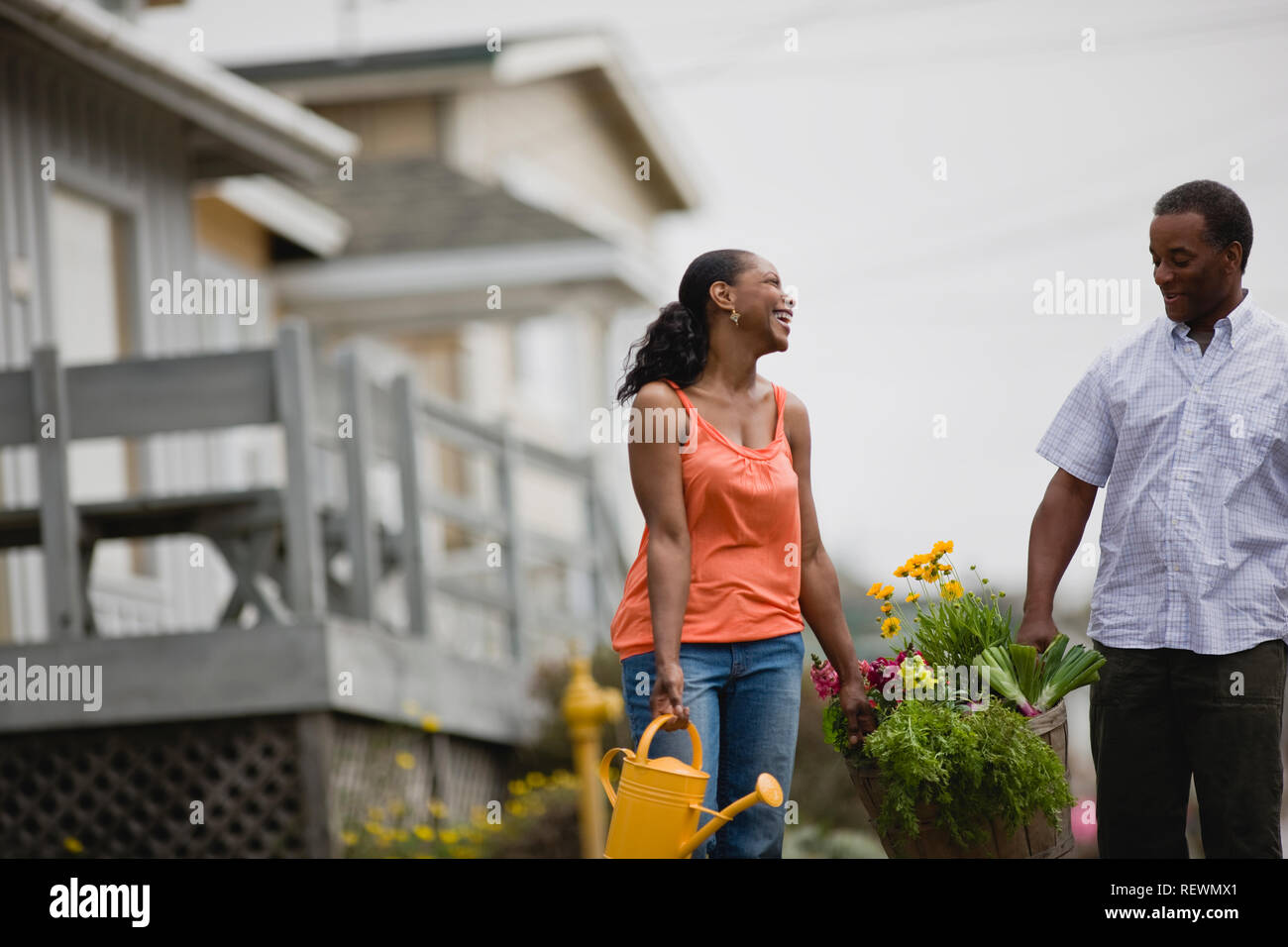 Happy mid-adult couple portant un panier en bois de plantes et d'arrosoir dans une rue de banlieue. Banque D'Images