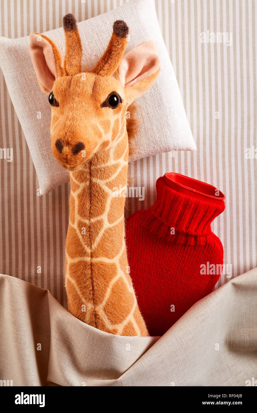 Peu de malades girafe en peluche à câliner dans un lit avec une bouteille d'eau chaude rouge vif pour garder au chaud dans un concept de soins pédiatriques Banque D'Images