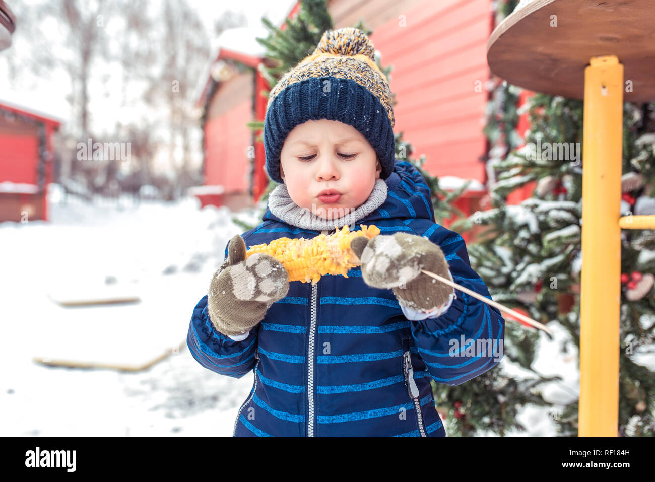 Un petit bébé garçon est en train de manger du maïs bouilli. Dans  l'après-midi d'hiver sur une marche. Dans un chapeau chaud avec une veste  bleue et verte des mitaines. Concept de