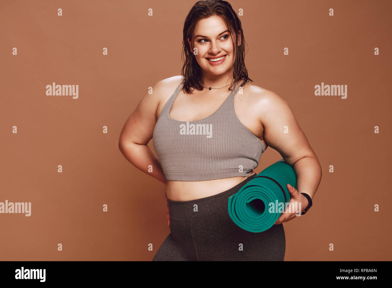 Jeune femme avec corps sain surdimensionné debout avec tapis de yoga en studio. modèle féminin de fitness en vêtements de sport après l'entraînement de yoga sur fond brun. Banque D'Images